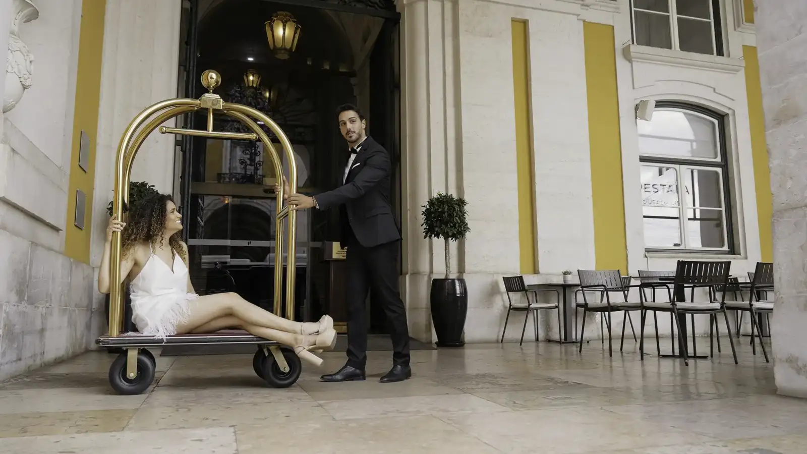 Un groom en costume pousse un chariot à bagages doré avec une femme assise.