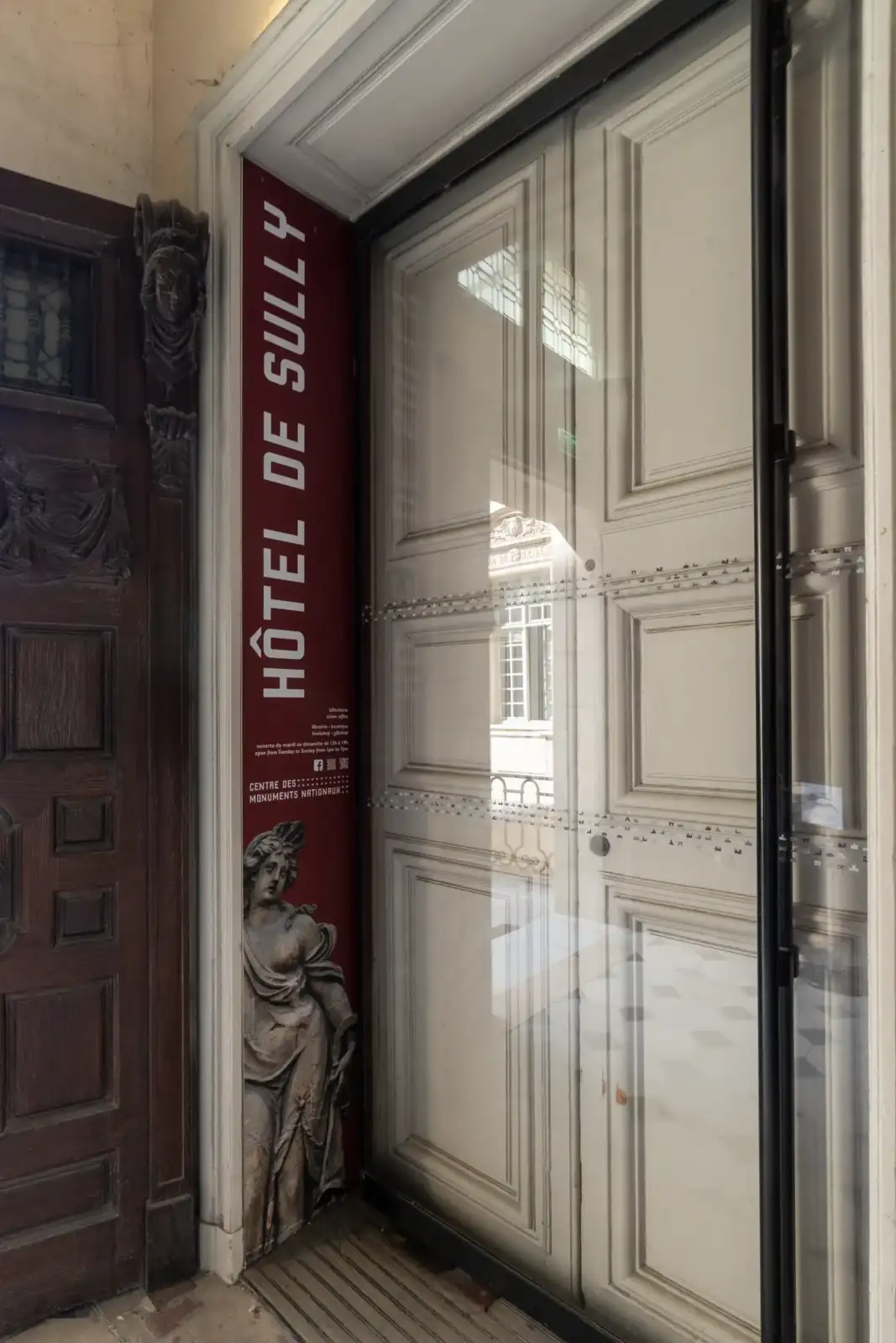 Une porte vitrée moderne encadrée par un panneau rouge indique l'entrée Hôtel de Sully.