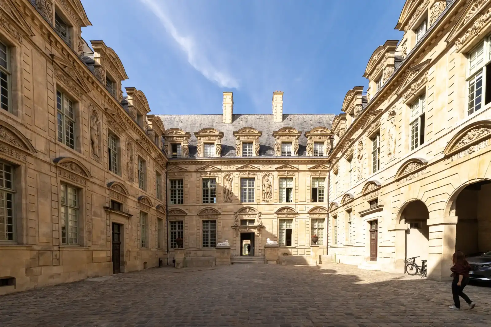 Une magnifique cour pavée entourée de façades en pierre sculptée sous un ciel bleu.