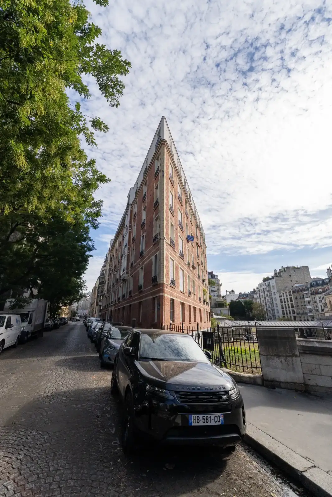 Vue en contre-plongée d’un immeuble triangulaire très étroit bordé de voitures stationnées.