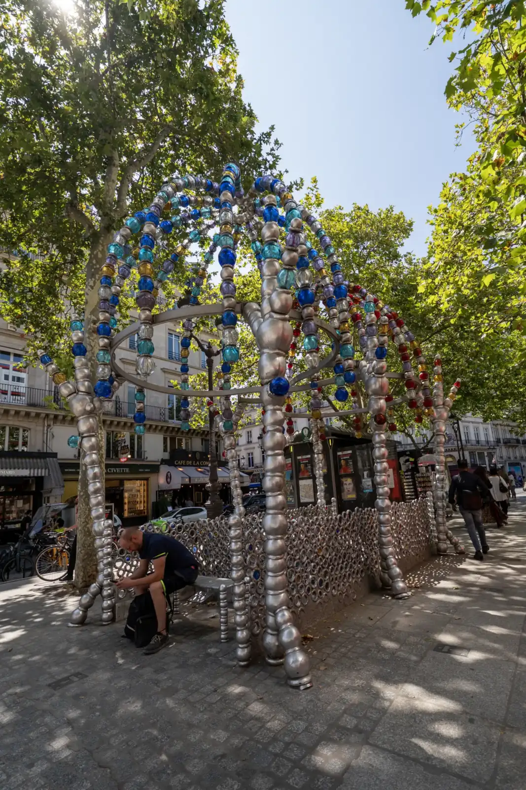 Une installation artistique métallique et colorée orne une rue animée de Paris sous les arbres.