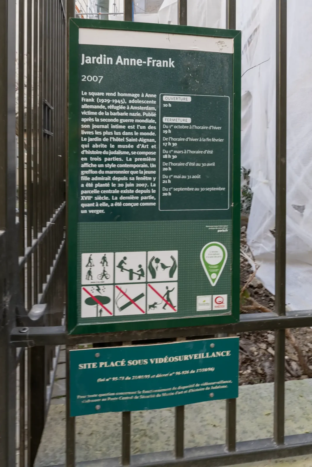 Panneau d'information vert sur une grille métallique devant le jardin Anne Frank à Paris.