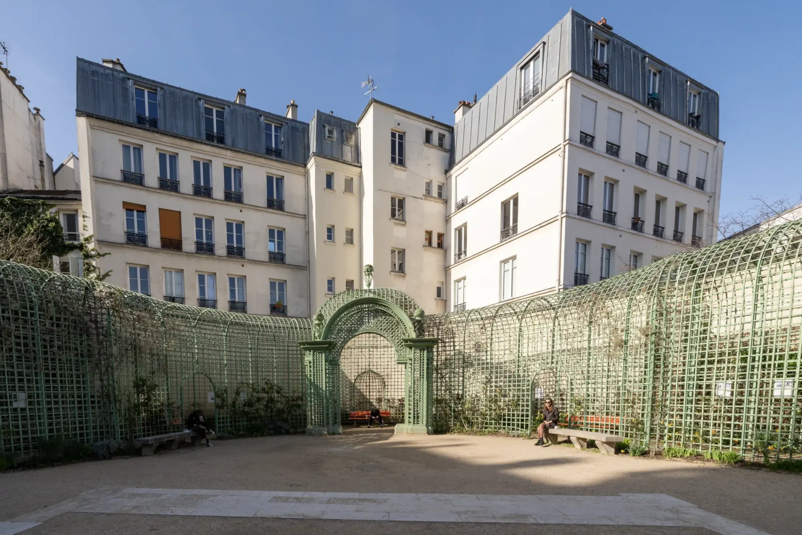 Une structure en treillage vert entoure une cour parisienne sous un ciel bleu clair.