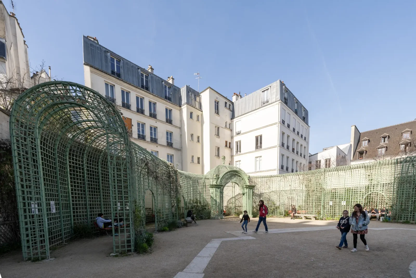 Des personnes se promènent dans le jardin Anne Frank entouré de treillages métalliques verts.