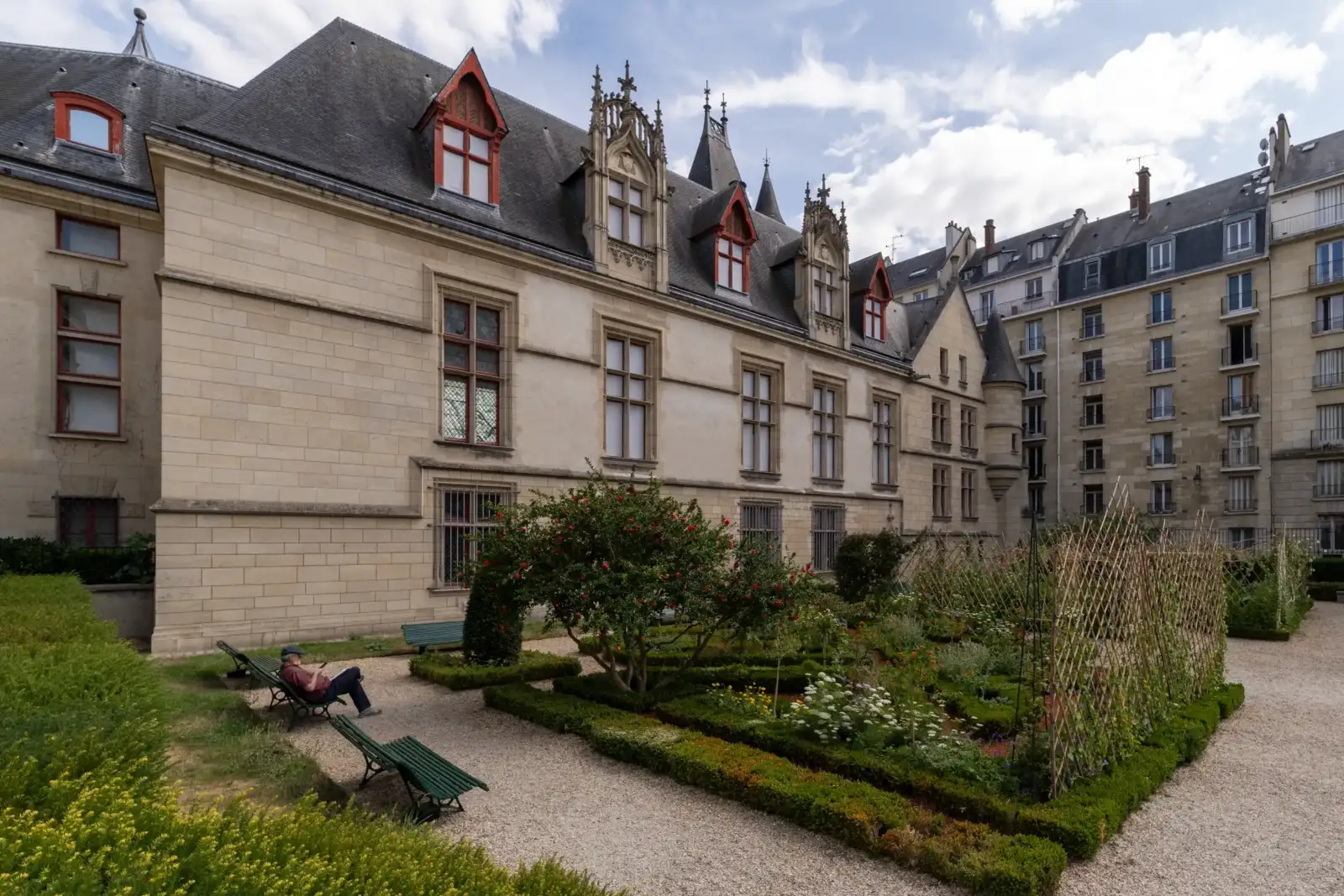 Une personne assise sur un banc lit dans le jardin de l'hôtel de Sens.