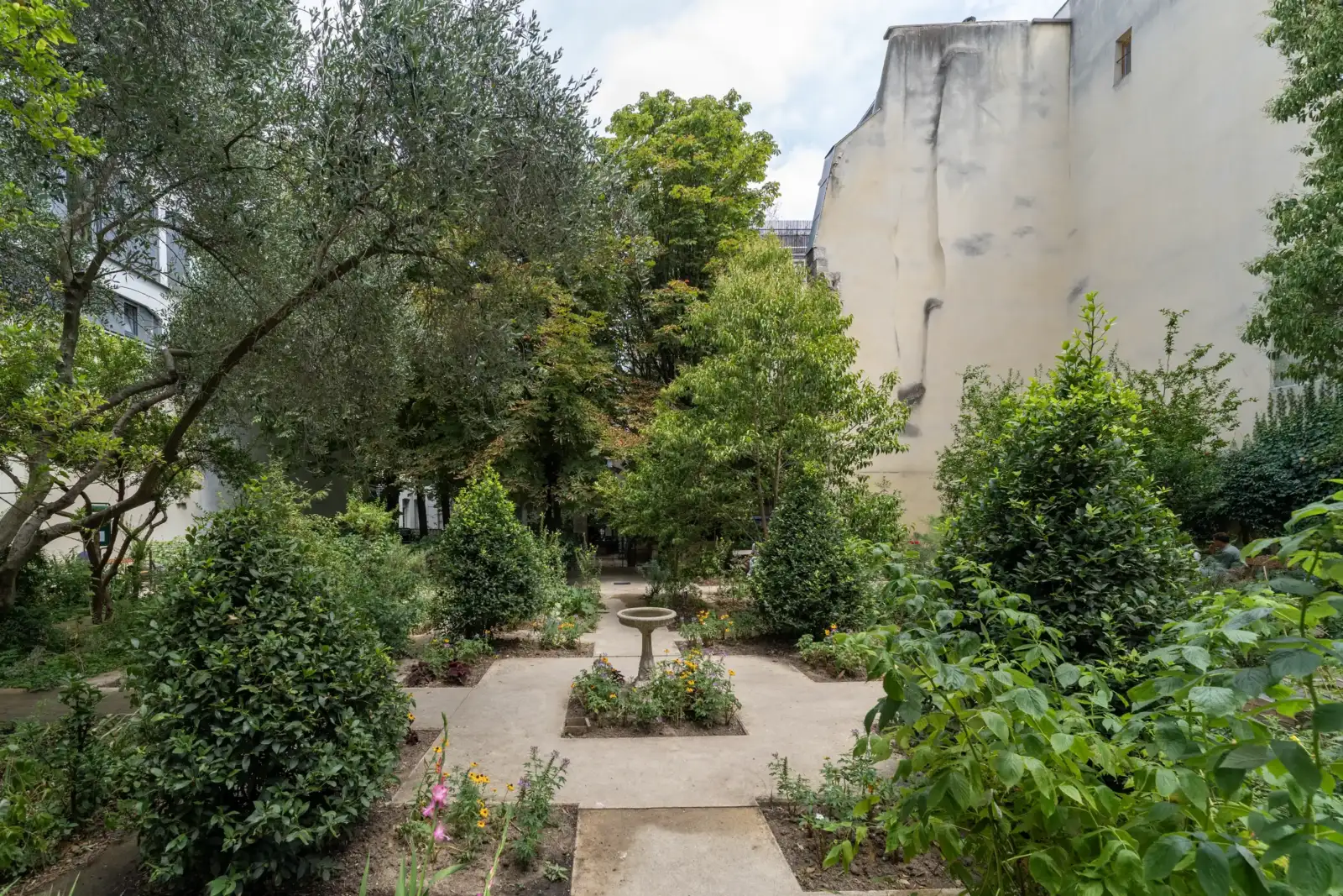 Une allée centrale traverse le paisible jardin parisien arboré et fleuri entre des façades d'immeubles.