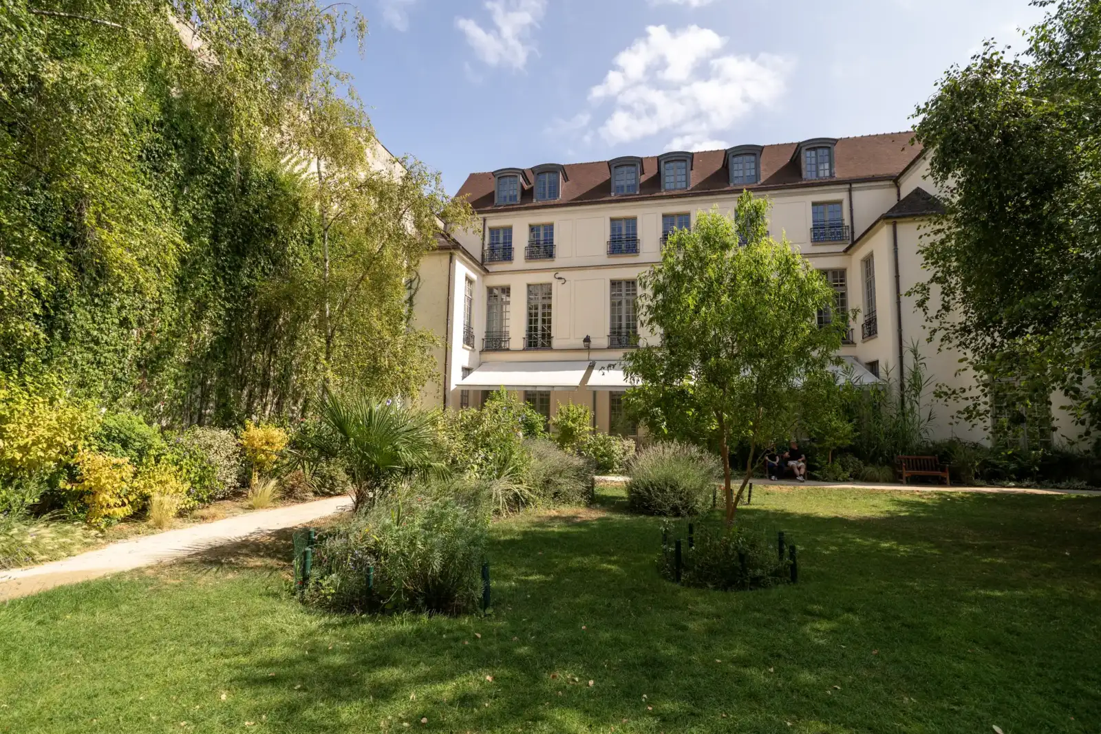 Facade claire et vegetalisée du jardin parisien Joseph Migneret, sous un ciel nuageux.