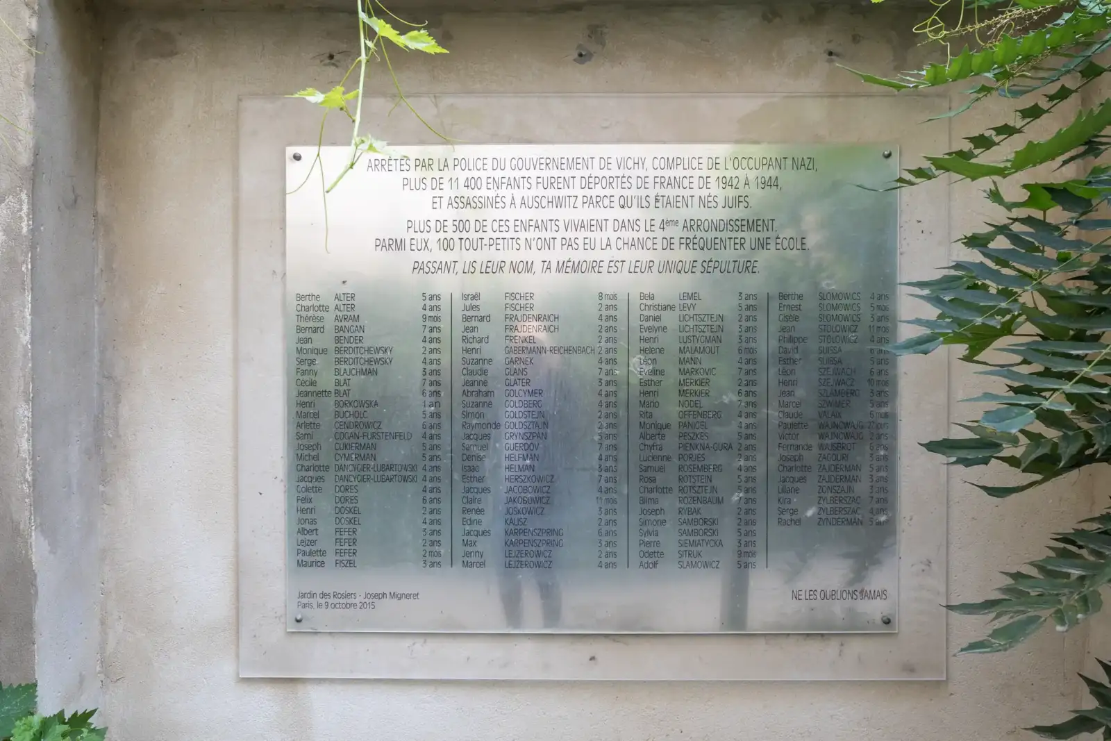 Plaque commémorative en verre avec des noms d enfants déportés, entourée de verdure.