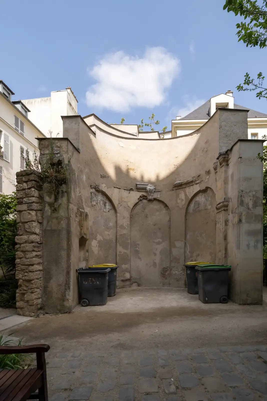 Un mur ancien en pierre présente des niches et abrite des poubelles.