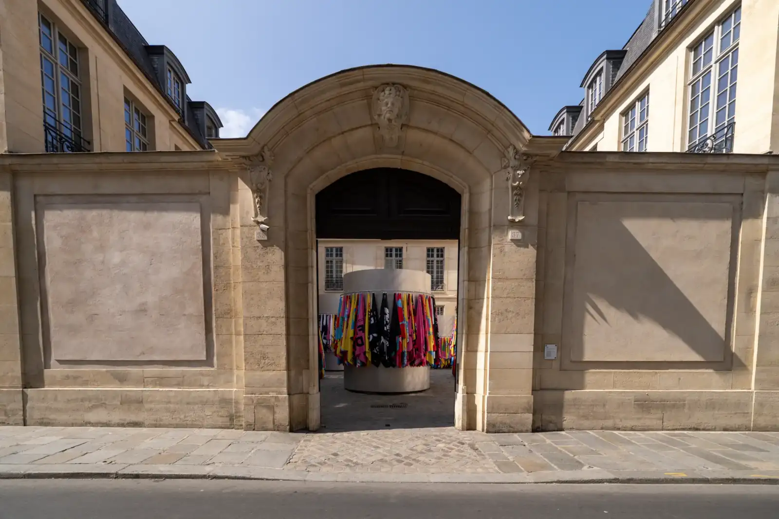 Un grand porche en pierre dévoile une exposition colorée de drapeaux accrochés dans une cour parisienne.