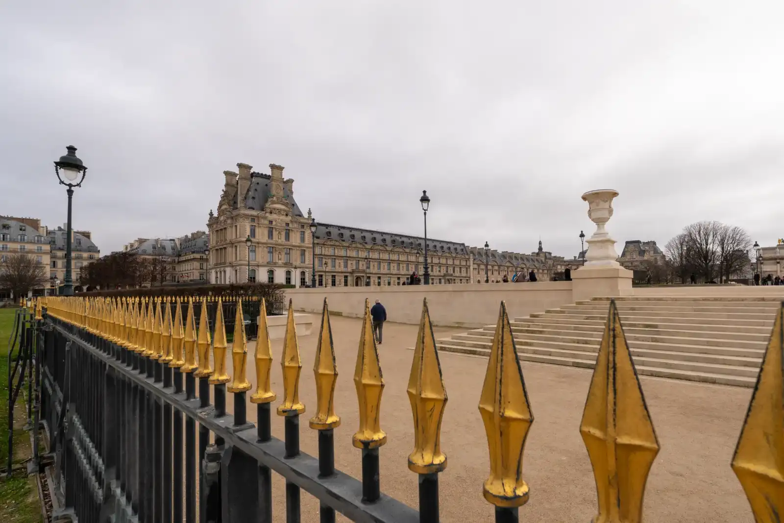 Une grille dorée borde une allée du jardin des Tuileries devant le palais Louvre.