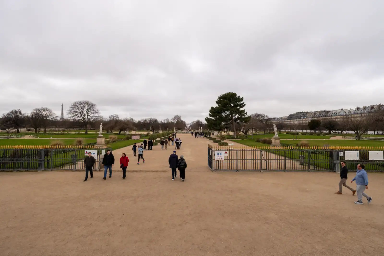 Des promeneurs traversent l'entrée du jardin des Tuileries sous un ciel parisien très nuageux.