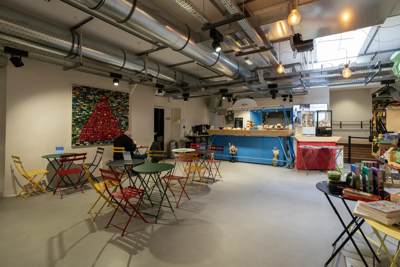 Un café moderne au plafond industriel avec des tables colorées et un comptoir bleu.