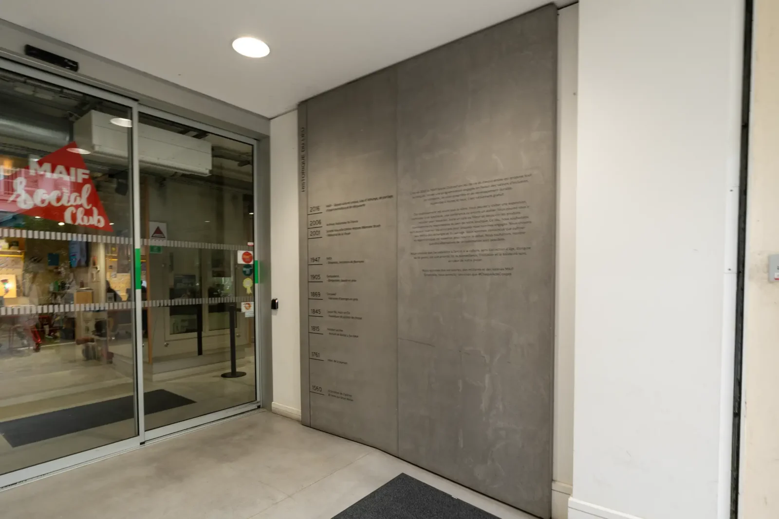 Entrée vitrée du MAIF Social Club à Paris avec un panneau historique mural gris.