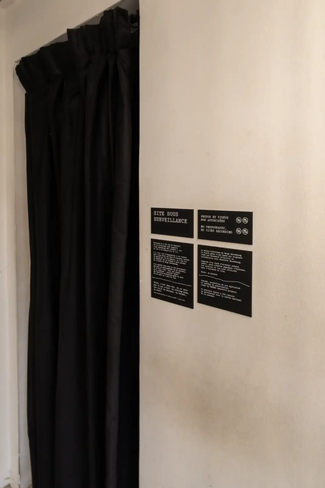 Un lourd rideau noir se dresse à côté de panneaux explicatifs sur un mur blanc.