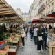 Des clients parcourent les étals d'un marché parisien traditionnel bordé d'immeubles en pierre.