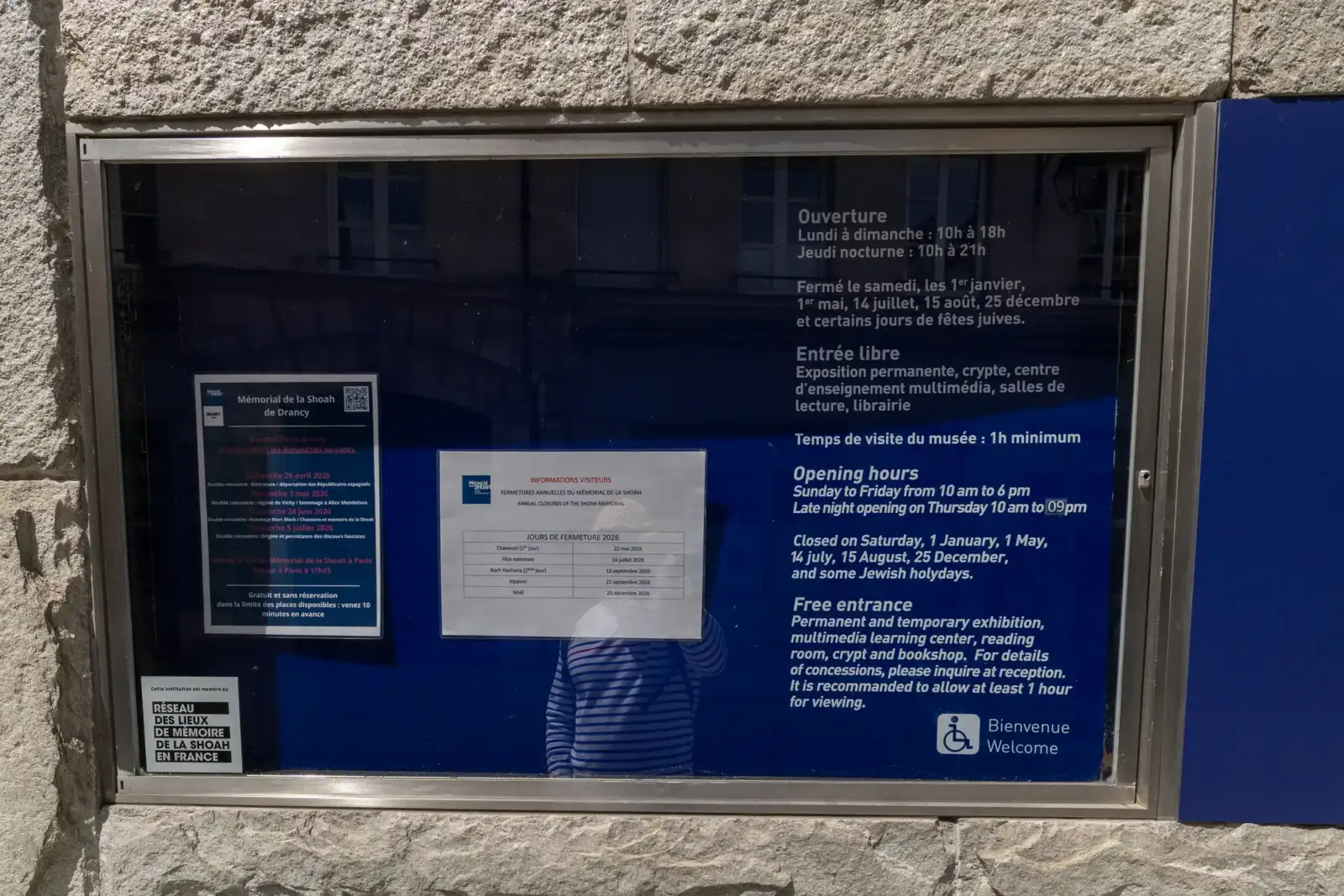 Un panneau d’information encastré dans un mur en pierre affiche les horaires en français et anglais.