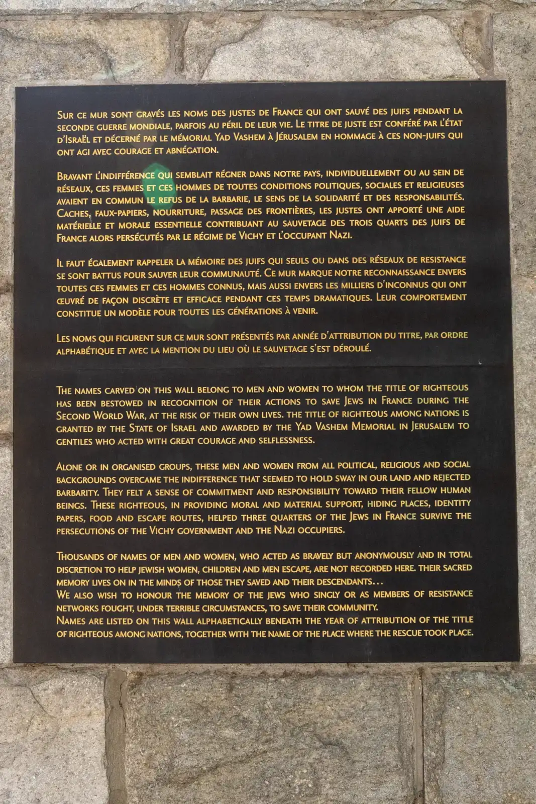 Une plaque explicative noire en français et anglais est fixée sur un mur de pierre.