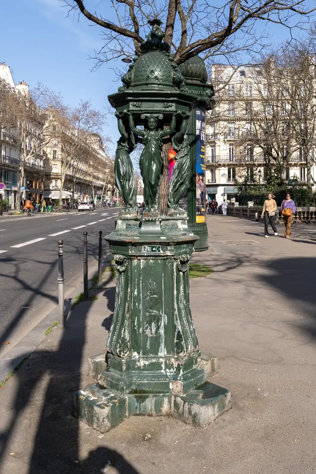 Une fontaine Wallace verte ornée de statues sur un trottoir parisien ensoleillé.