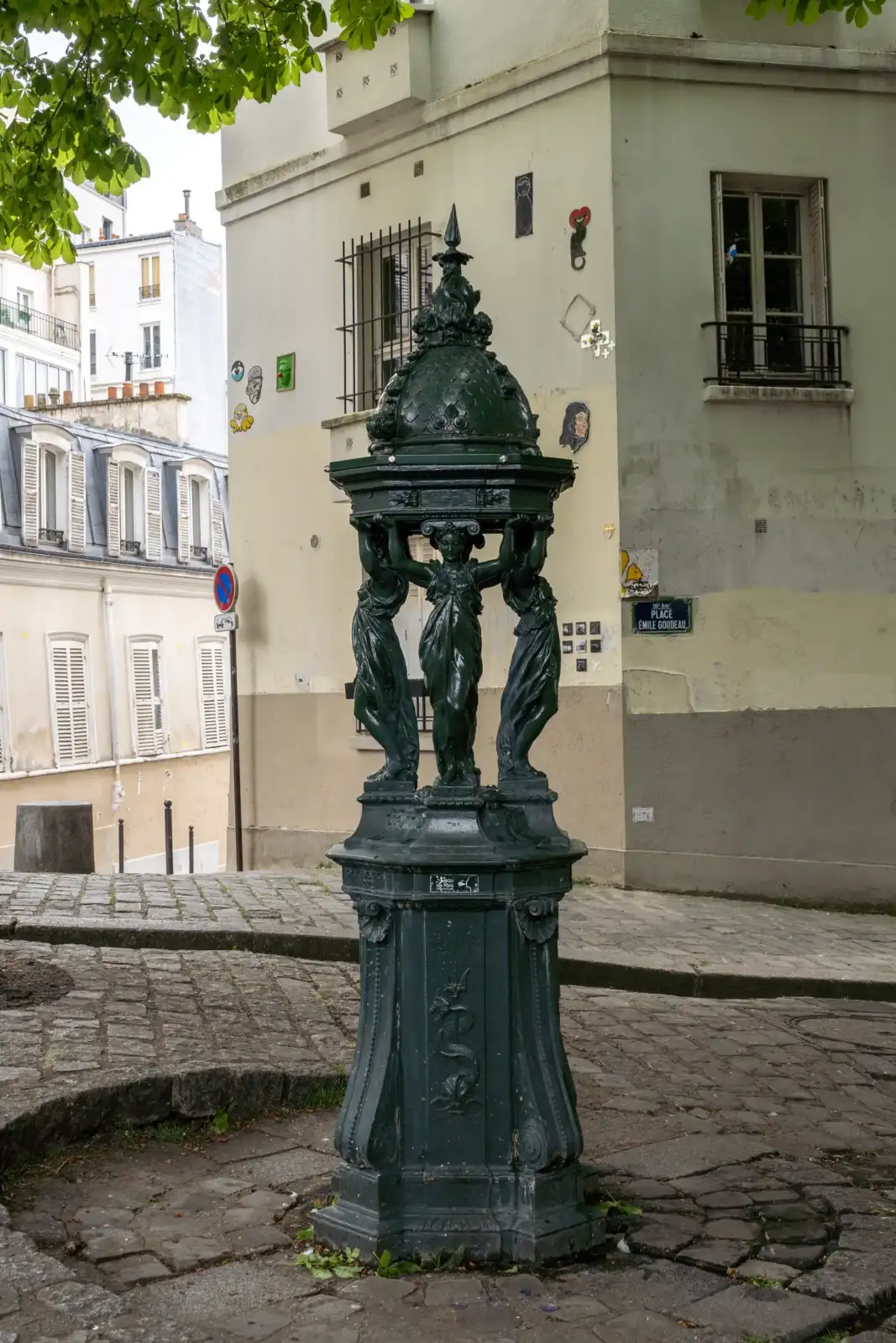 Une fontaine Wallace verte sur une place pavée parisienne devant des immeubles anciens.