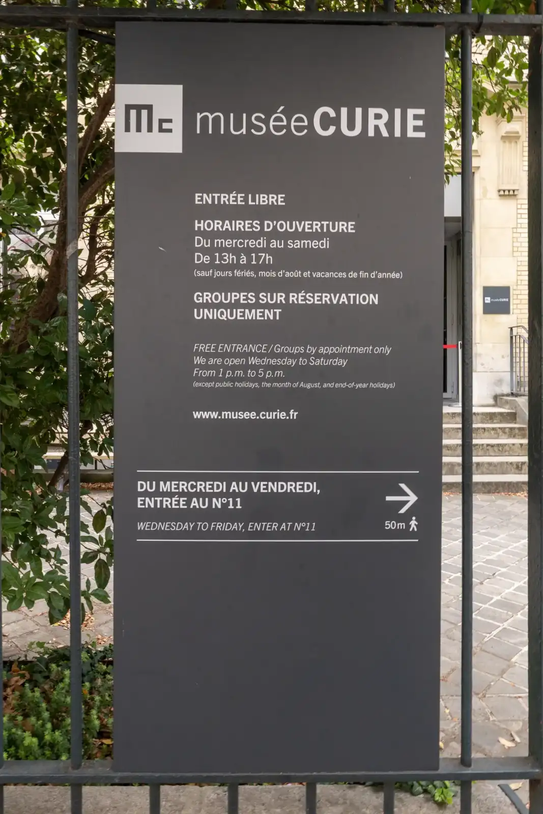Un panneau d'affichage gris indique les horaires d'ouverture du musée Curie à Paris.