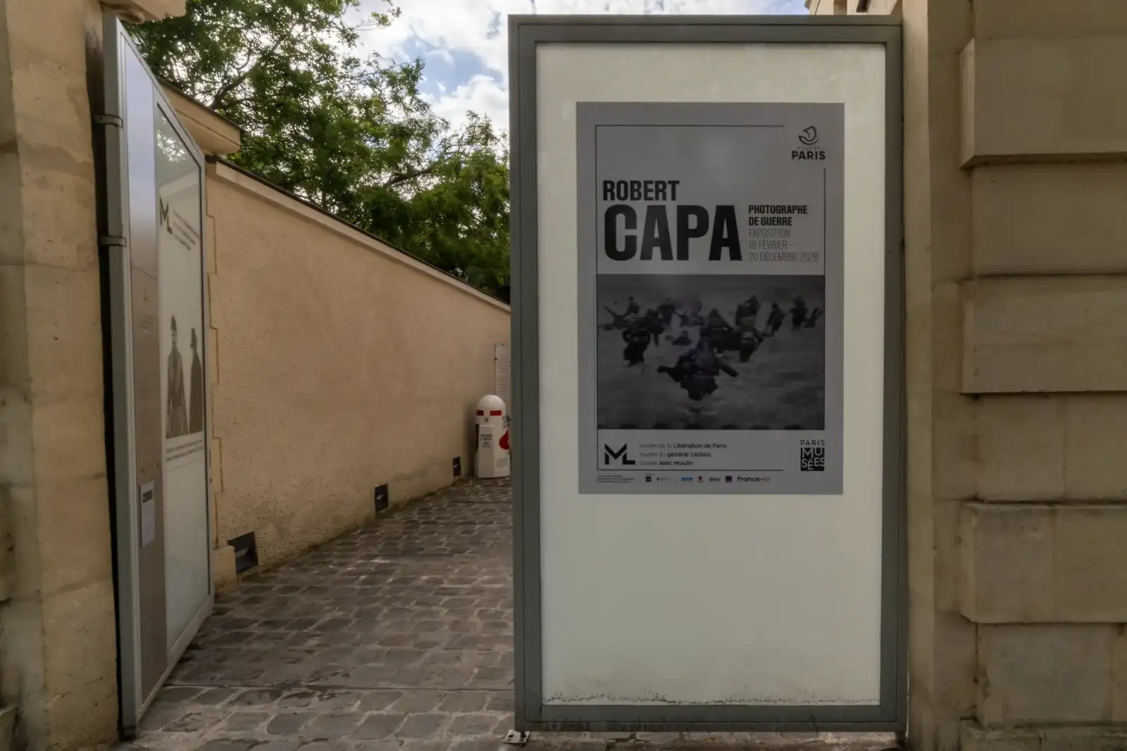 Panneau d'affichage annonçant une exposition sur Robert Capa devant une allée pavée.