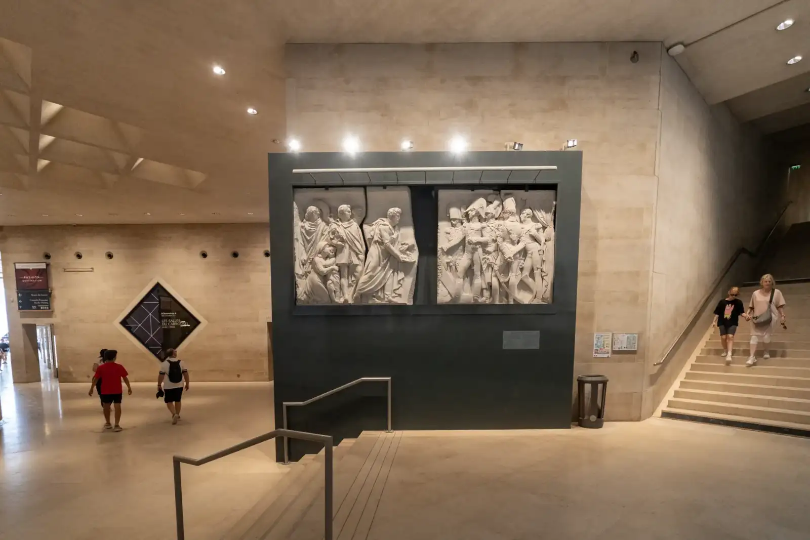 Des bas-reliefs sculptés sont exposés sur un mur dans un hall moderne du Louvre.