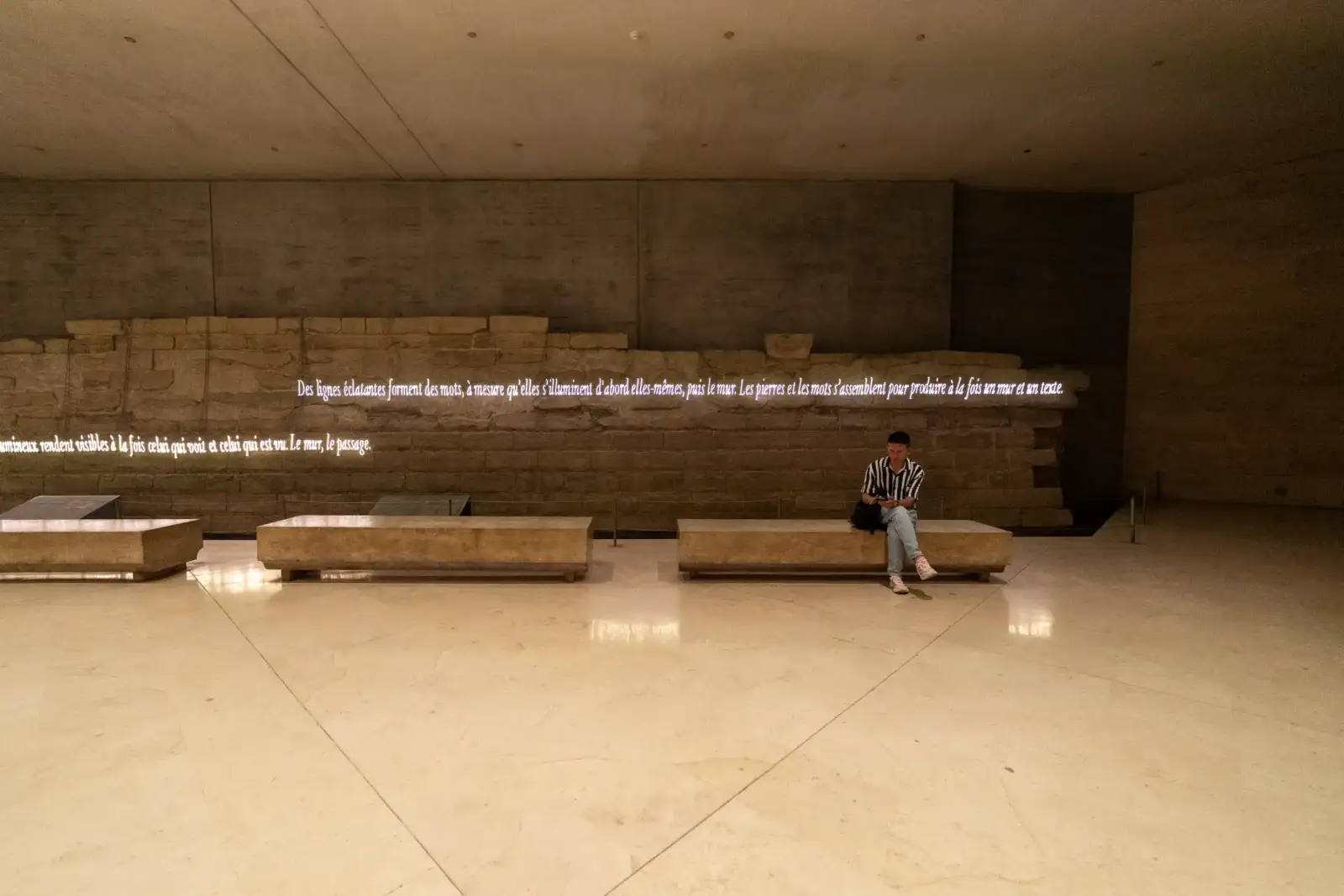 Un homme assis sur un banc devant un ancien mur en pierre illuminé de texte.