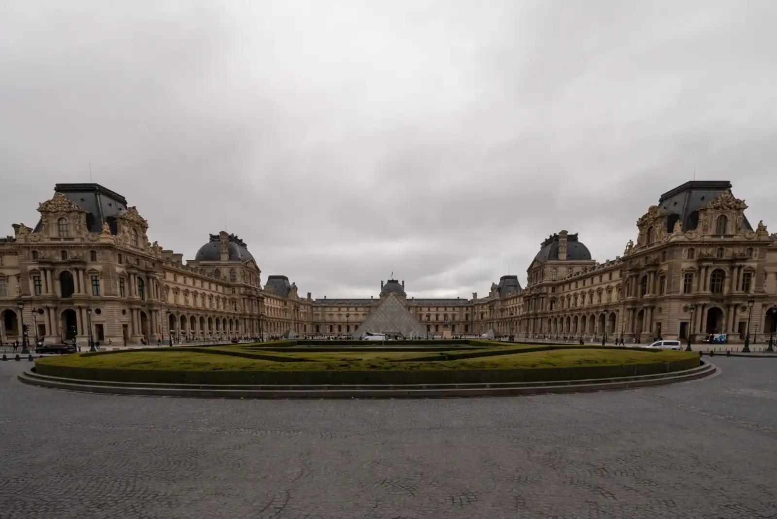 La façade historique du musée du Louvre entoure la célèbre pyramide sous un ciel gris.