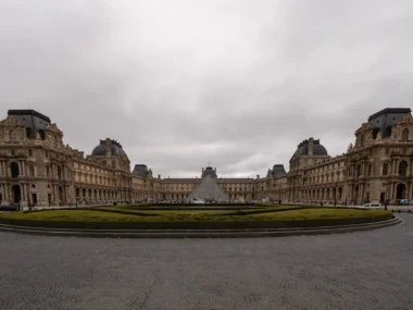 La façade historique du musée du Louvre entoure la célèbre pyramide sous un ciel gris.