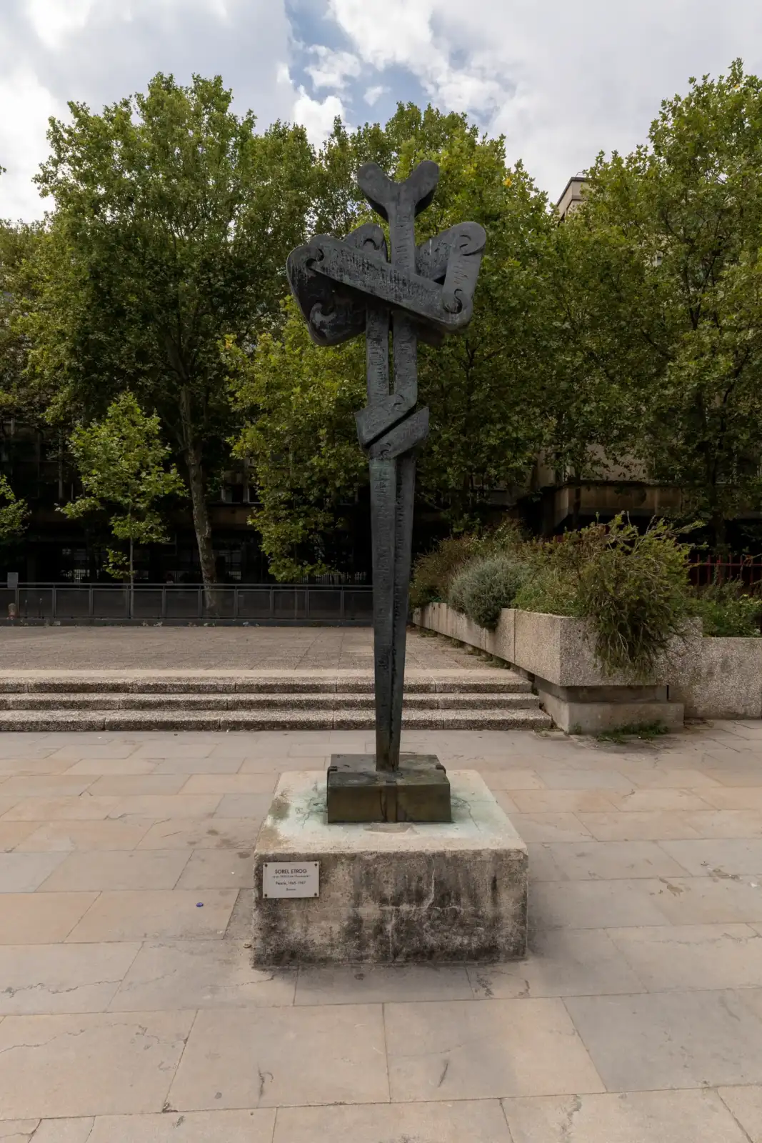 Une sculpture en bronze sombre se dresse sur un socle en pierre pavée en plein air.