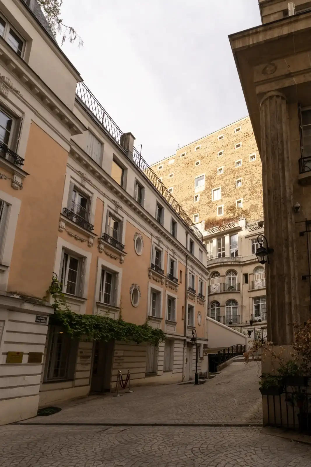 Vue de rue parisienne pavée encadrée par des immeubles anciens aux façades claires.