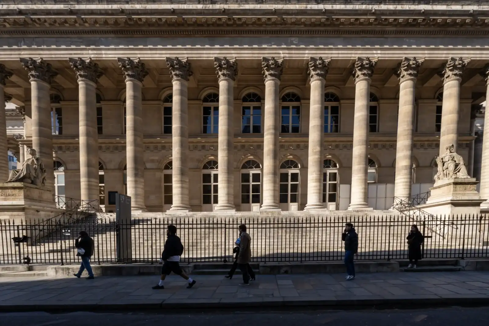 Vue de face du palais Brongniart à Paris avec ses colonnes classiques et des passants.