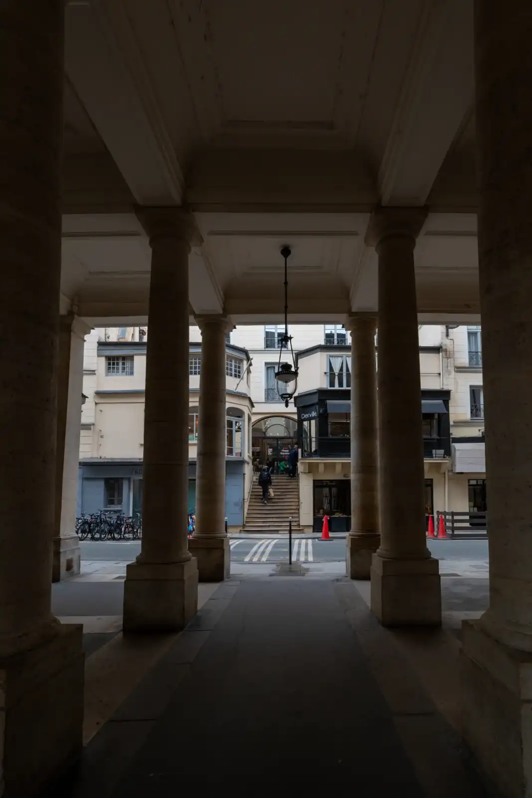 Vue sous les arcades du Palais-Royal montrant une façade parisienne et des escaliers extérieurs.
