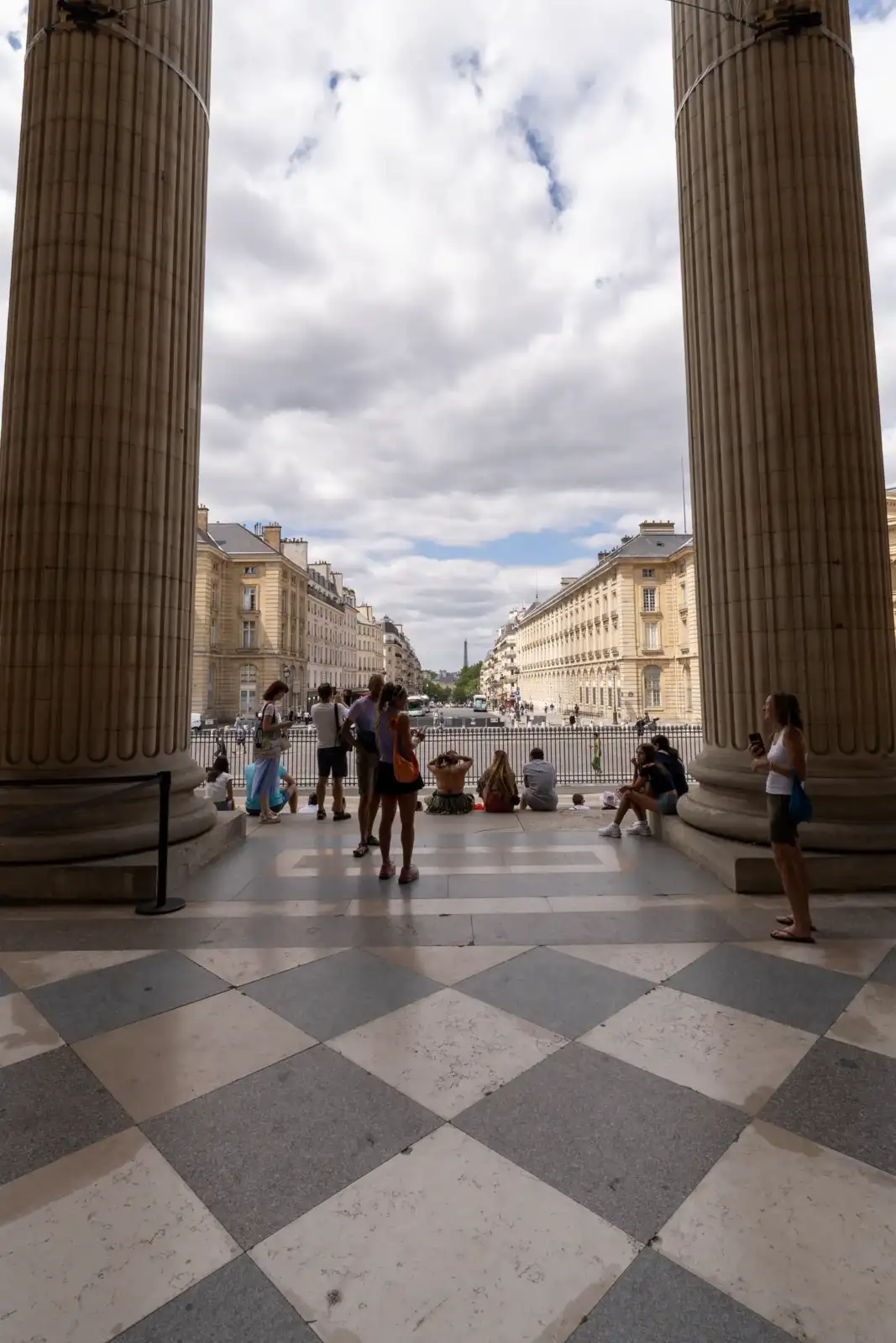 Vue encadrée par de hautes colonnes montrant des touristes devant des immeubles parisiens.