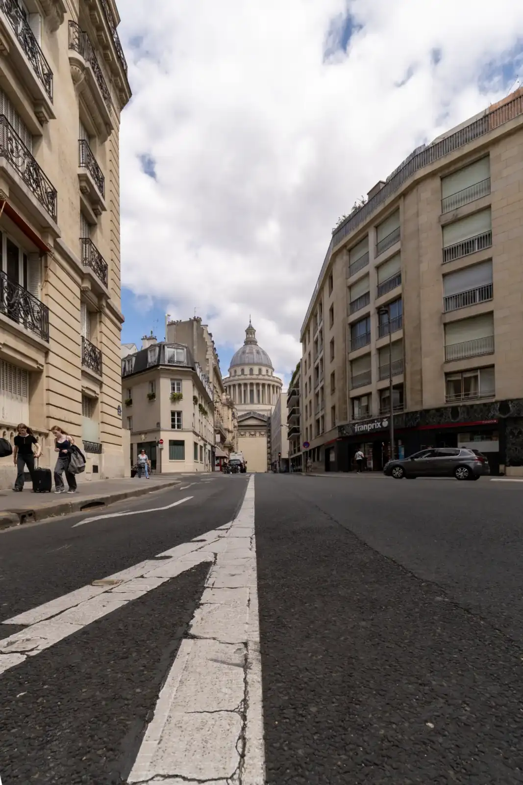 Vue depuis une rue parisienne montrant le majestueux Panthéon sous un ciel nuageux.