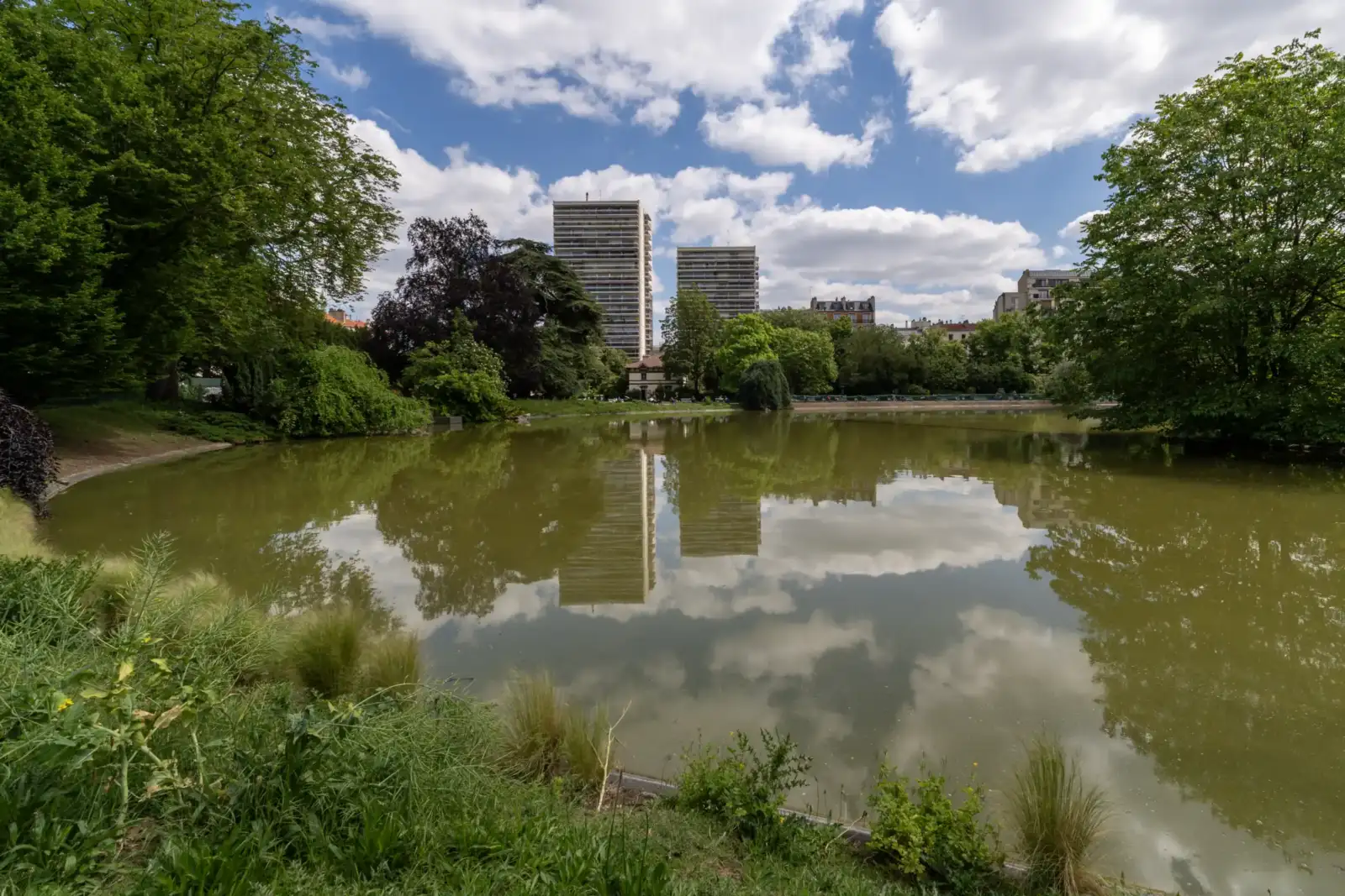 Le lac du parc Montsouris reflète des immeubles parisiens sous un ciel nuageux.