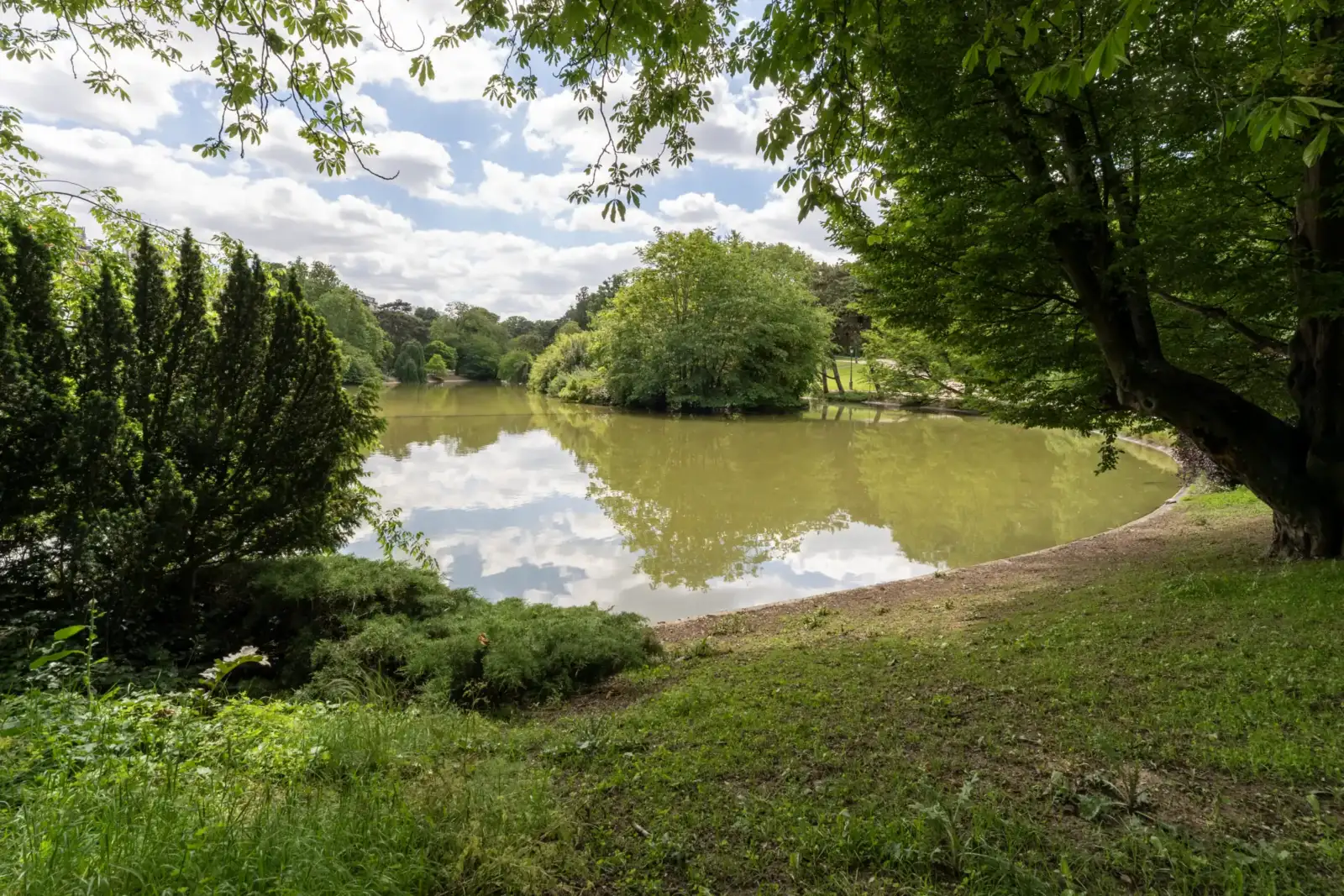 Vue paisible sur le lac verdoyant du parc Montsouris sous un ciel nuageux.