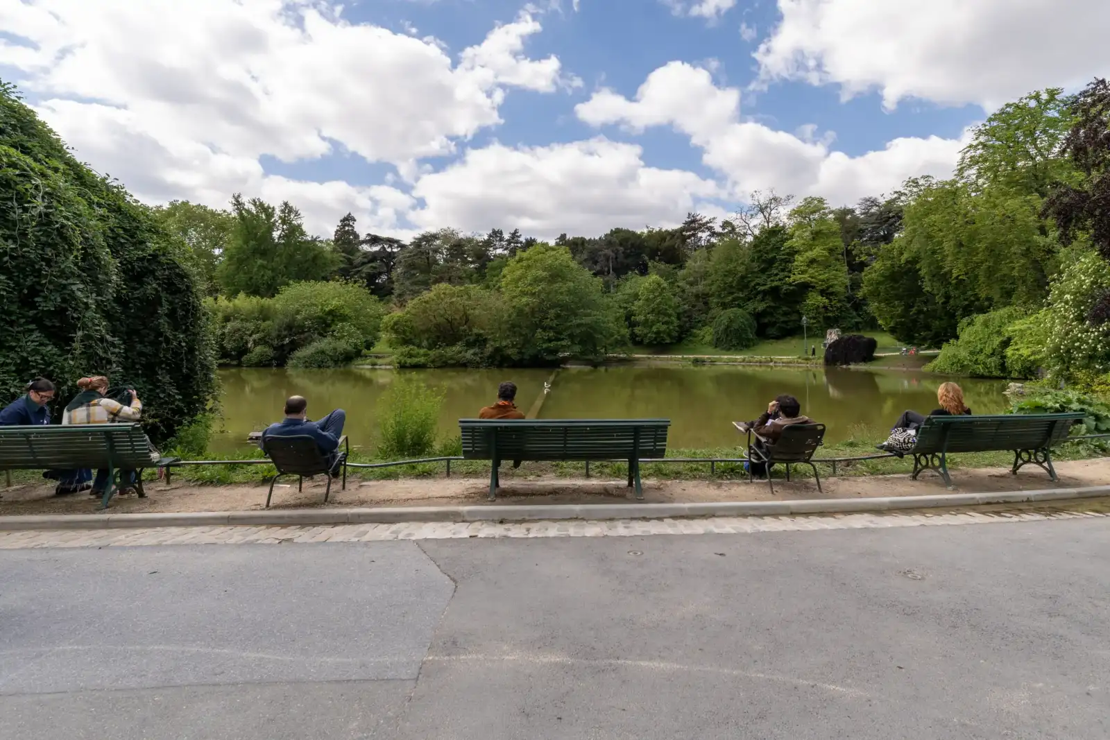 Des personnes sont assises sur des bancs face à un étang au parc Montsouris.