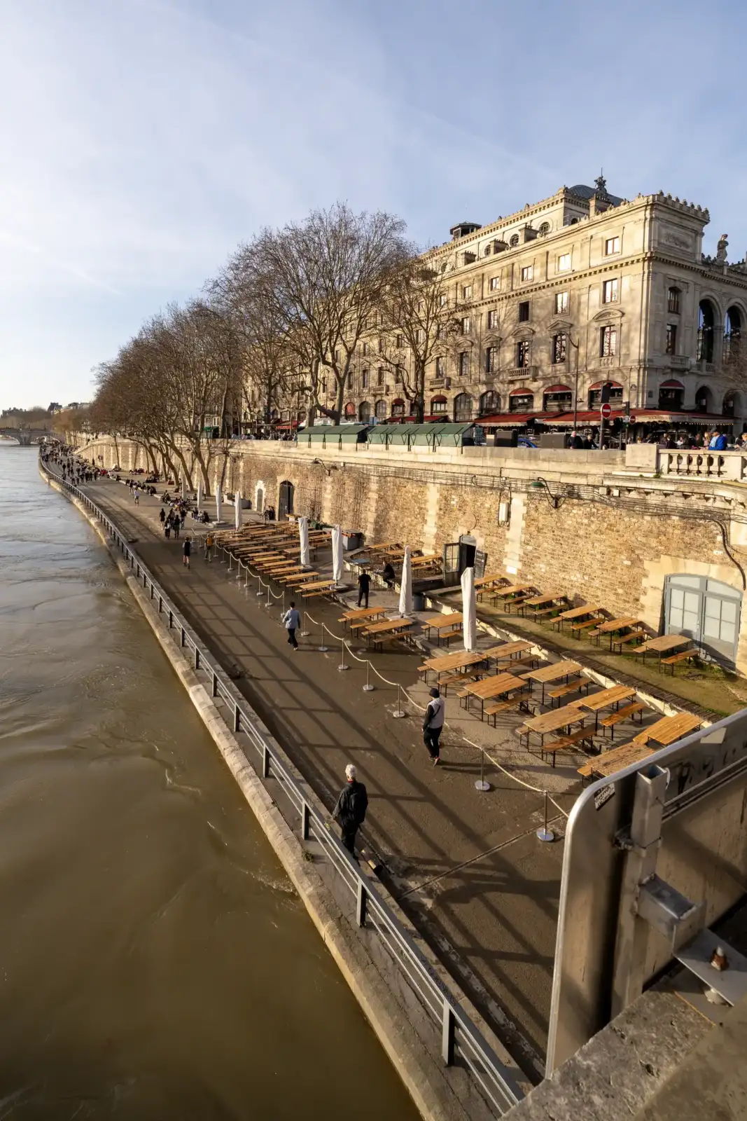 Promenade ensoleillée le long de la Seine avec des terrasses vides et des arbres nus.