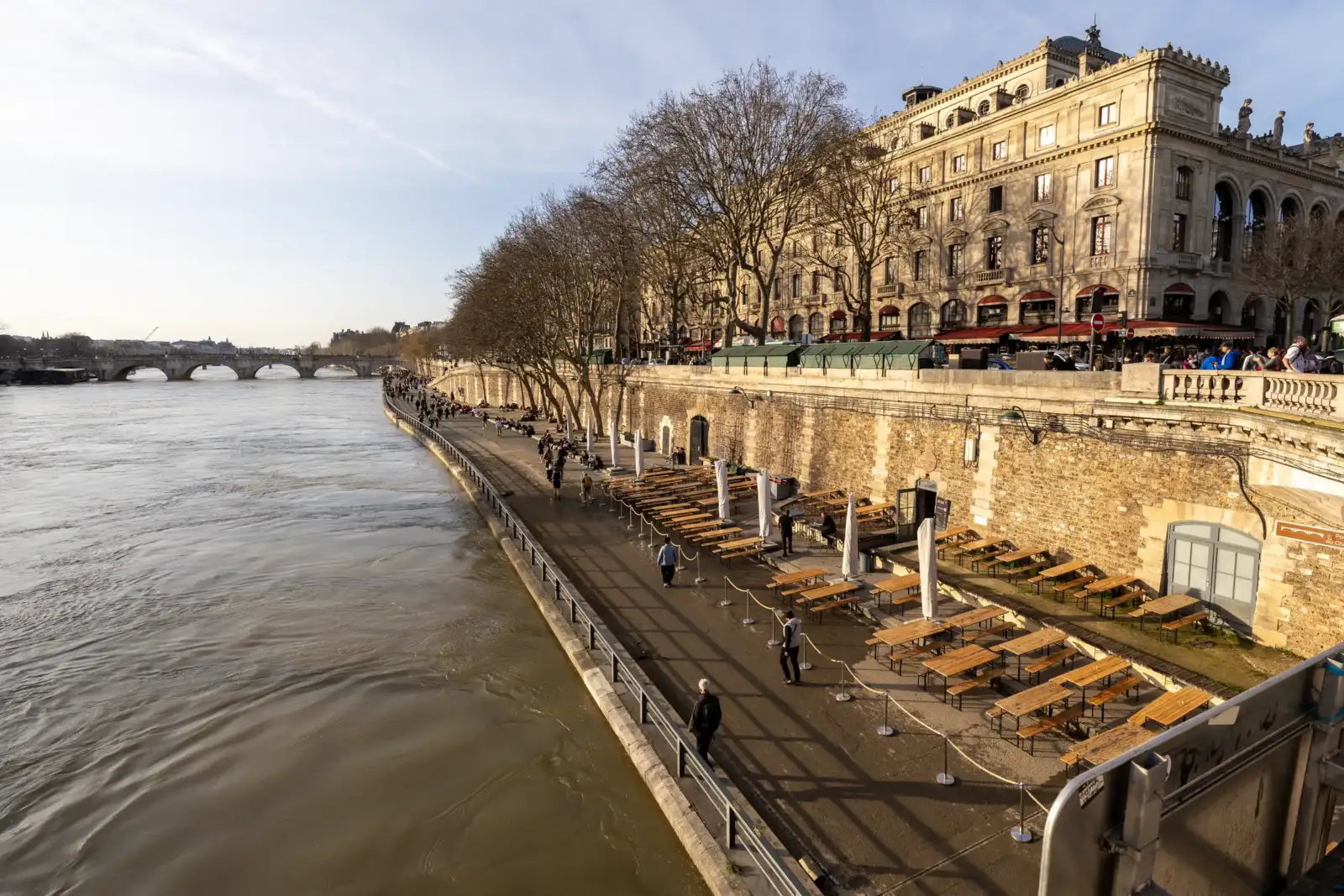 Promenade ensoleillée le long de la Seine avec des terrasses vides et des arbres nus.