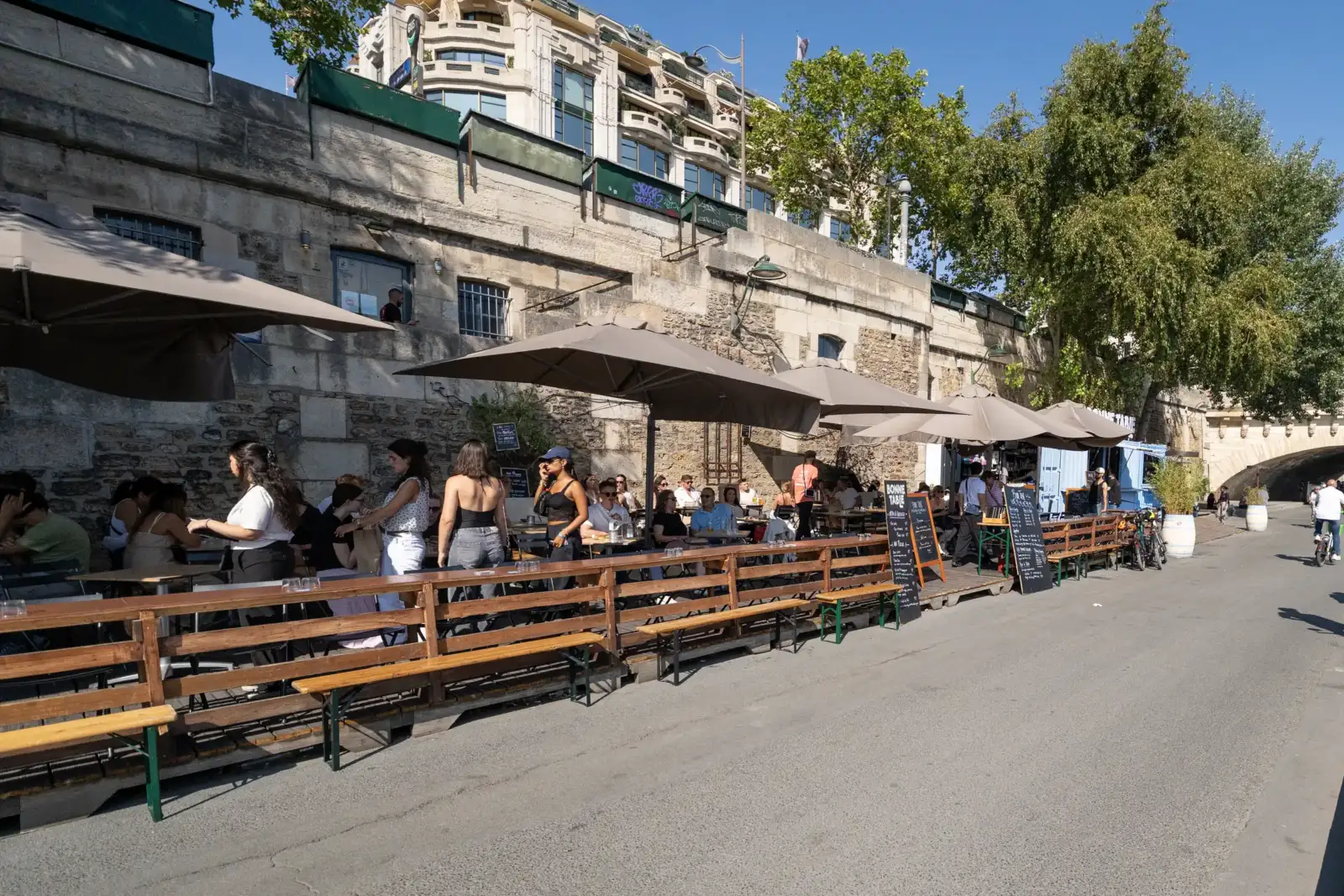 Une terrasse de café animée ensoleillée avec de grands parasols gris le long d'un quai parisien.