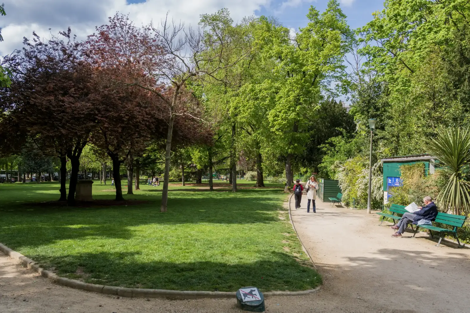 Des promeneurs profitent d'un parc parisien verdoyant sous un ciel bleu parsemé de nuages.