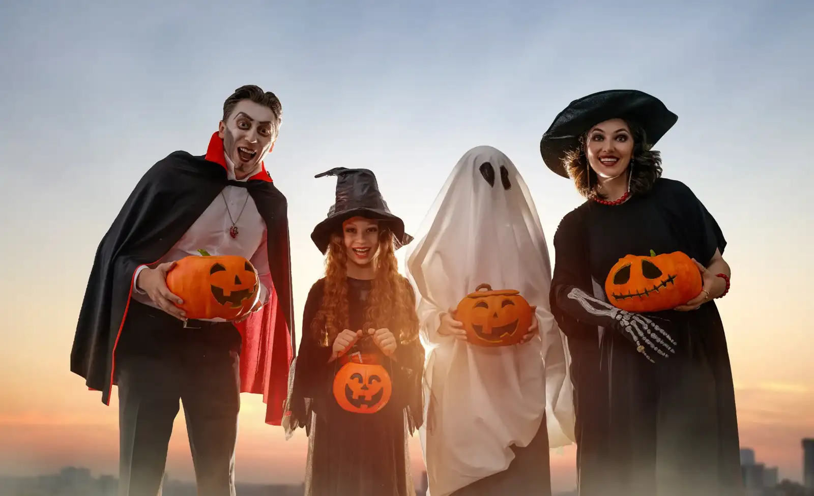 Une famille déguisée pour Halloween tient des citrouilles sculptées devant un ciel crépusculaire.