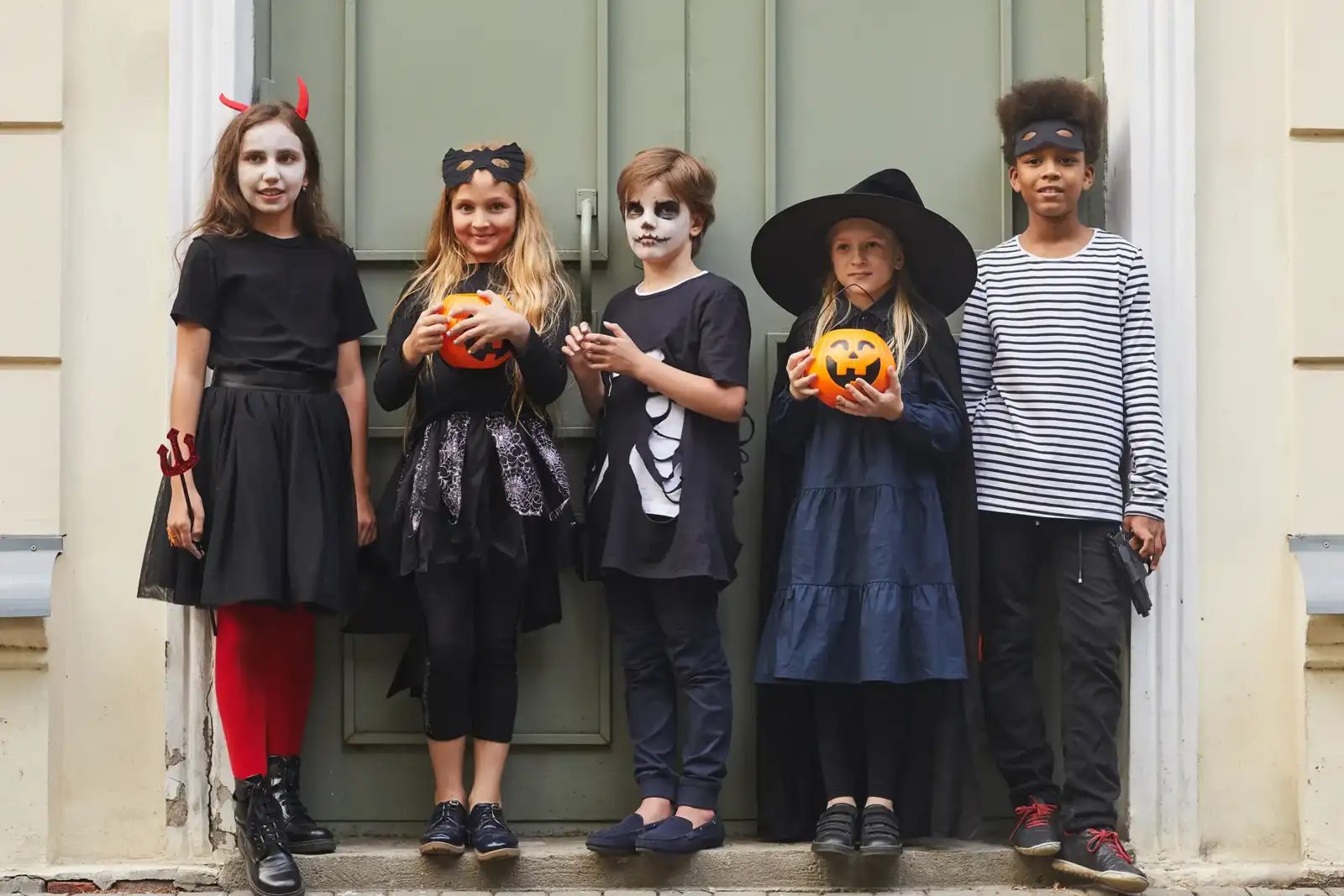 Cinq enfants déguisés pour Halloween posent devant une porte verte avec des seaux citrouilles.