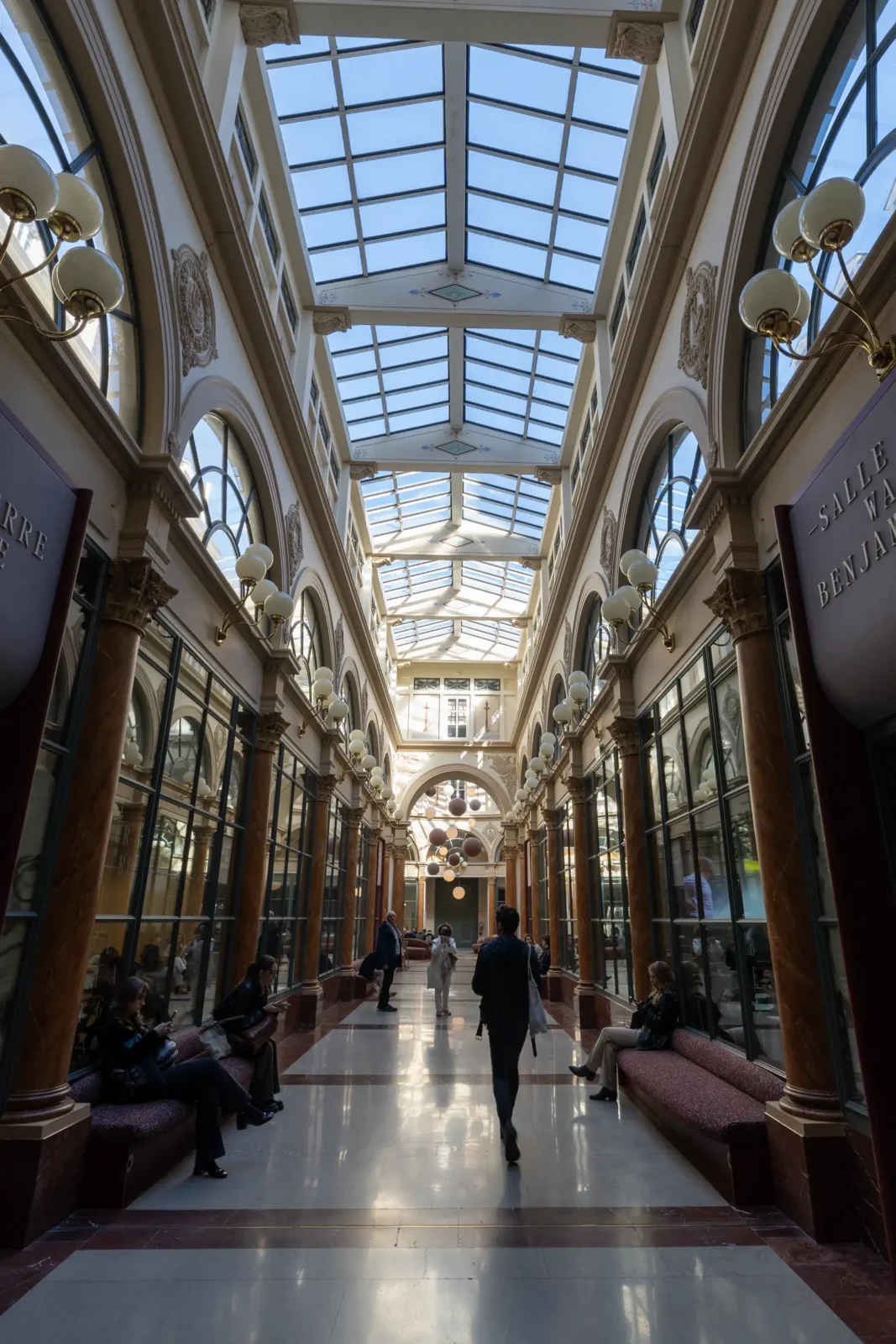 Vue intérieure du Passage Colbert à Paris avec une verrière et des passants assis.