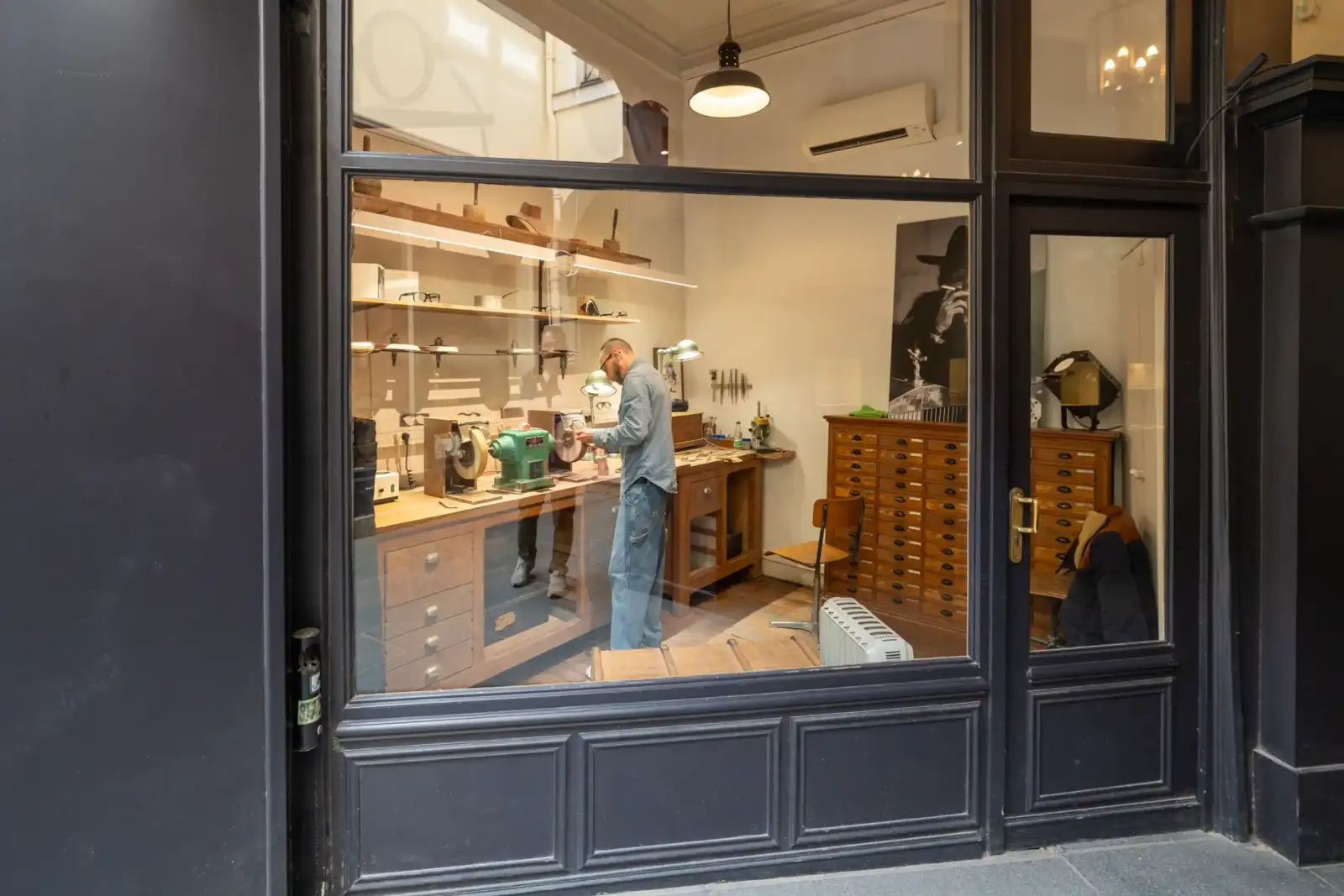 Un artisan travaille dans son atelier parisien visible à travers une vitrine sombre.