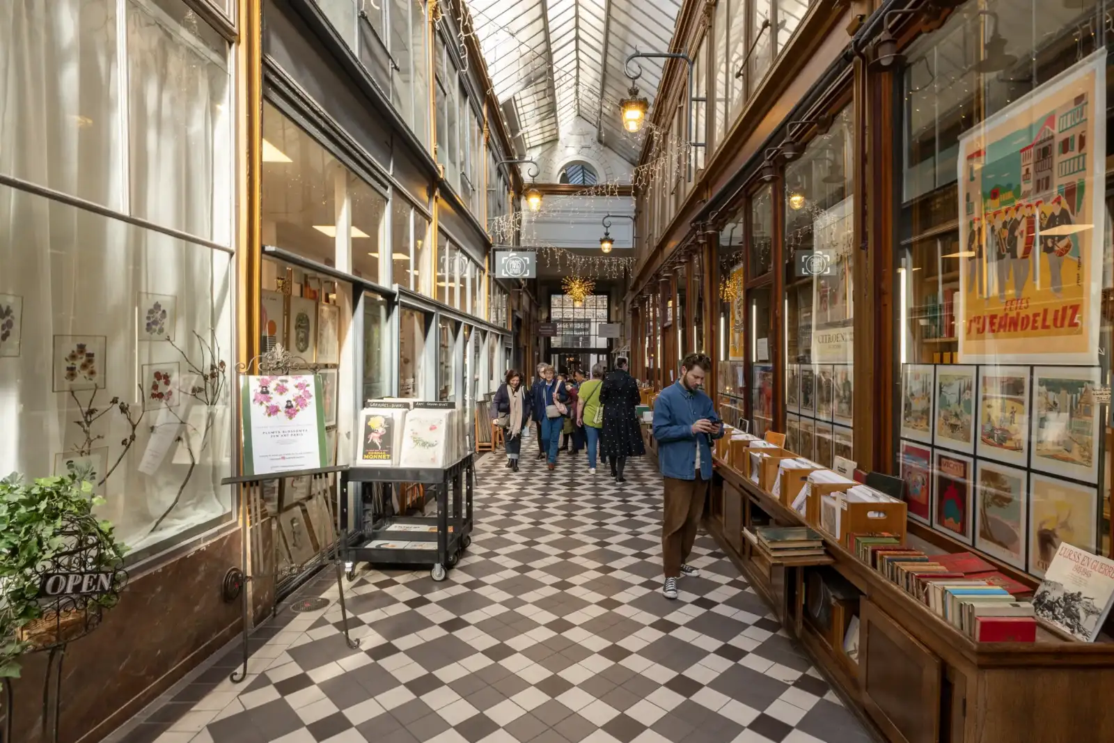 Un homme debout et des passants déambulent dans le passage Jouffroy à Paris sous une verrière.