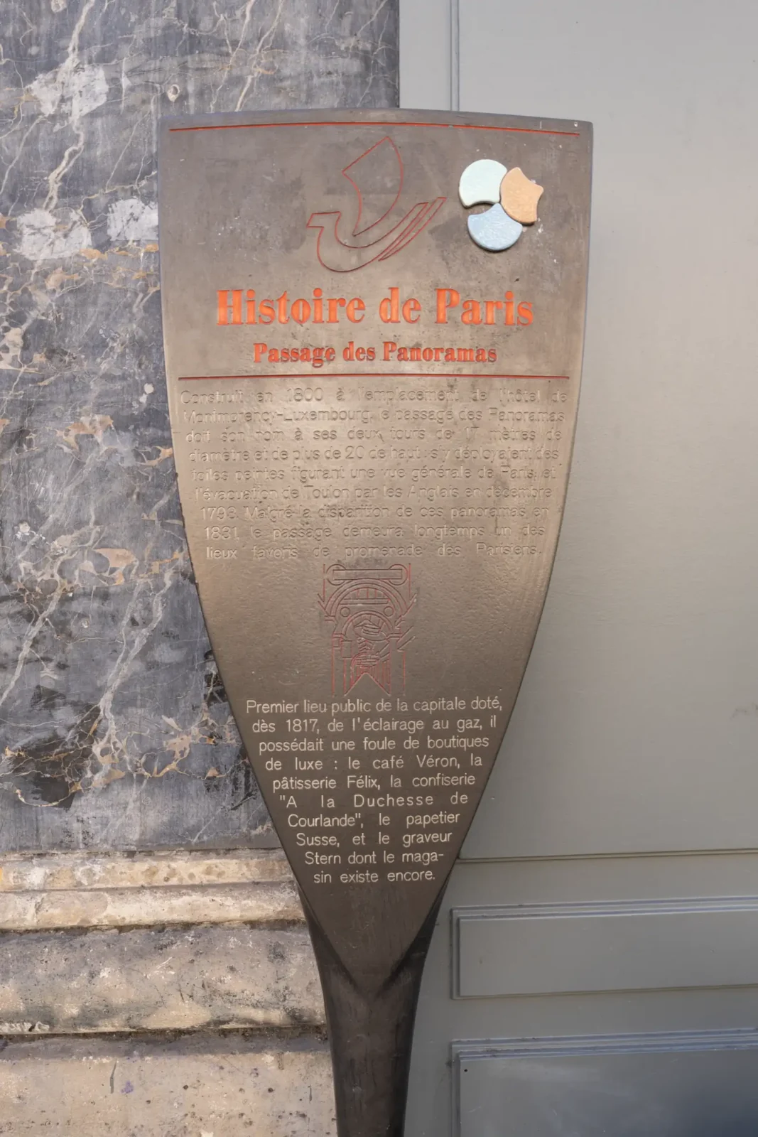 Plaque historique en métal sur un mur racontant l'histoire du Passage des Panoramas parisien.