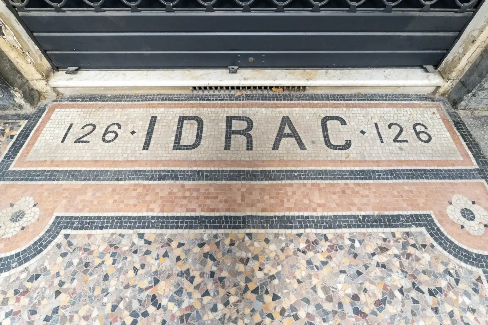Une mosaïque au sol affiche le texte 126 IDRAC 126 devant une porte métallique.